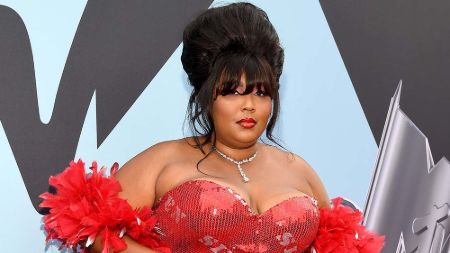 lizzo stands on the red carpet of VMA awards on a shiny red gown with a red furry scarf. he hair did in a high bun 